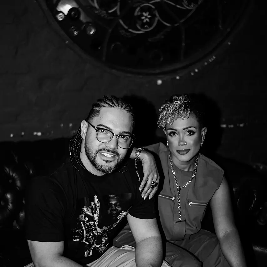 Carol e Daniel, as duas vozes da Black SP, sentados lado a lado num sofá de couro, embaixo de um vitral