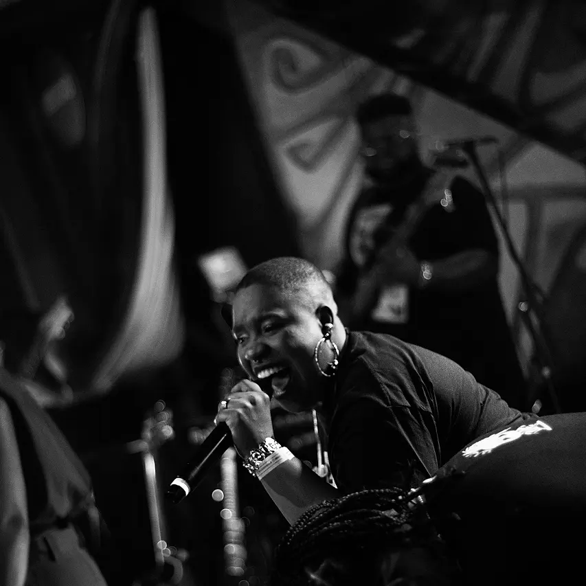Cantora da Black SP rindo ao microfone no palco do Black Music Live, com a banda tocando atrás
